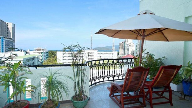 A balcony with a view of the city, two wooden chairs, a large umbrella, and potted plants. - La Suisse Hotel (Nha Trang)
