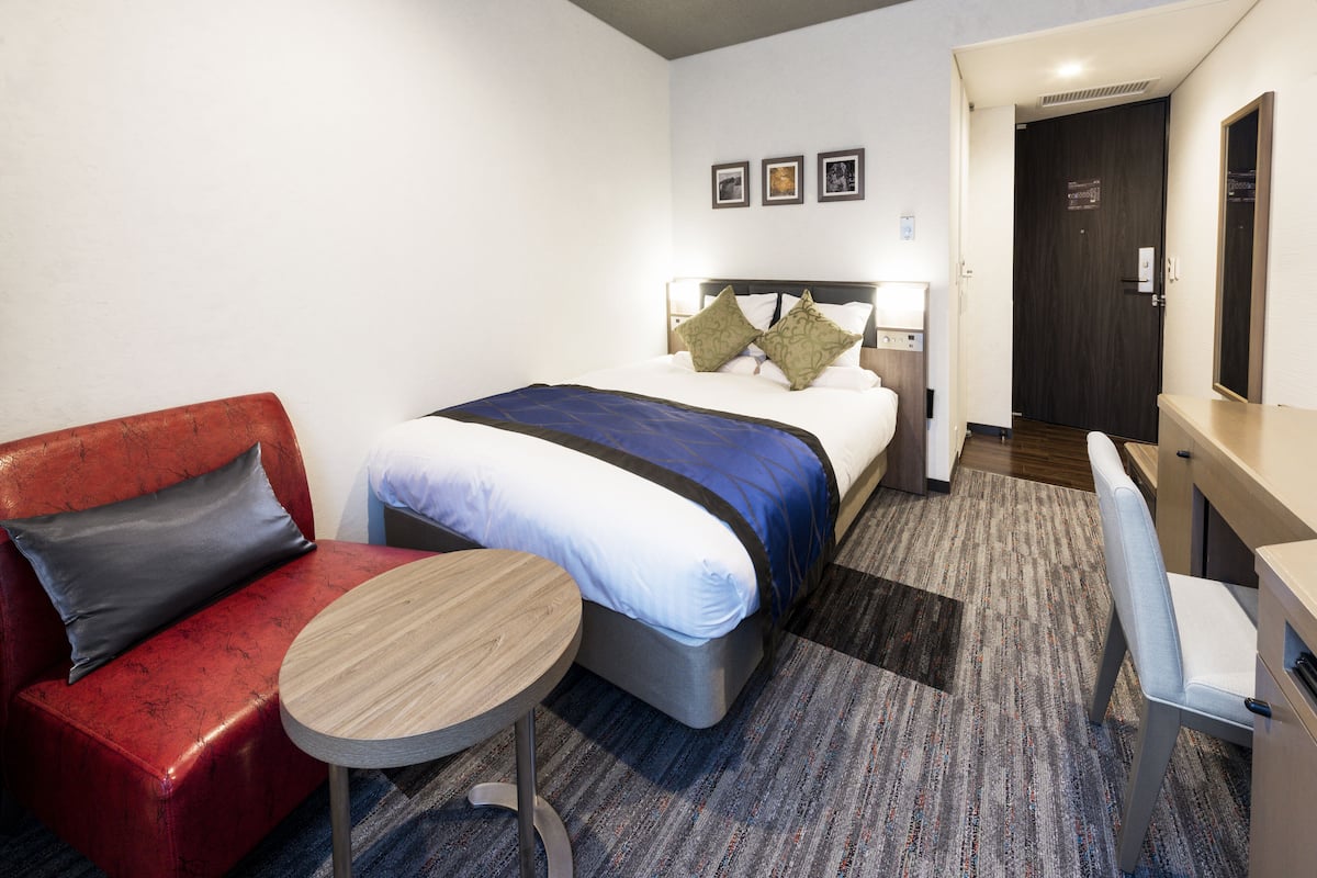 A hotel room with a bed, a red sofa, a small round table, and a desk with a chair.