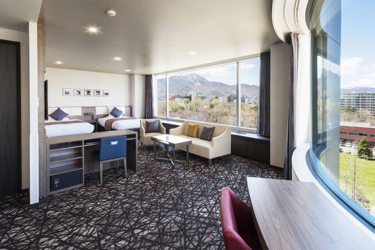A hotel room with a bed, a sofa, a table, and a large window offering a mountain view.