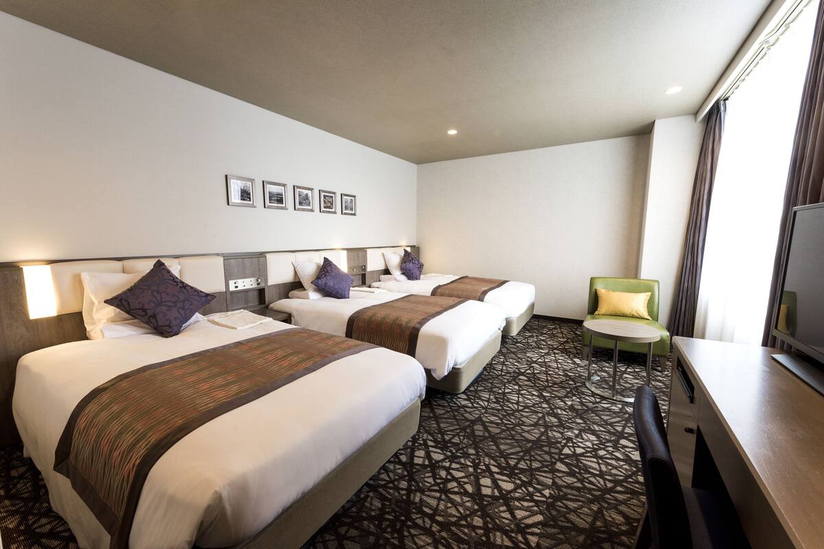 A hotel room with three beds, a small round table, a green armchair, and a TV.