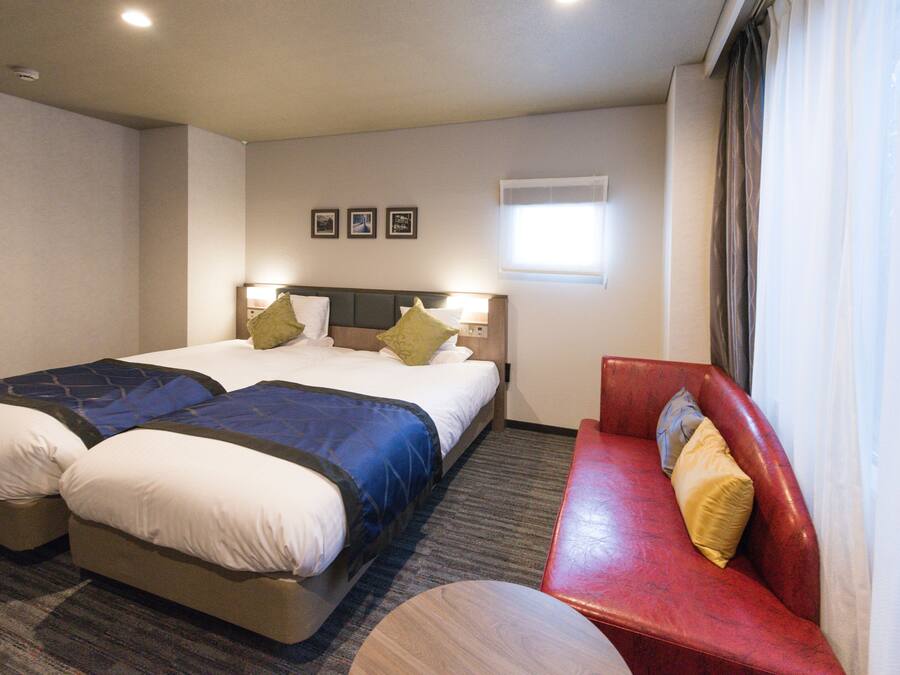 A hotel room with a double bed, a red sofa, a small round table, and a window with curtains.