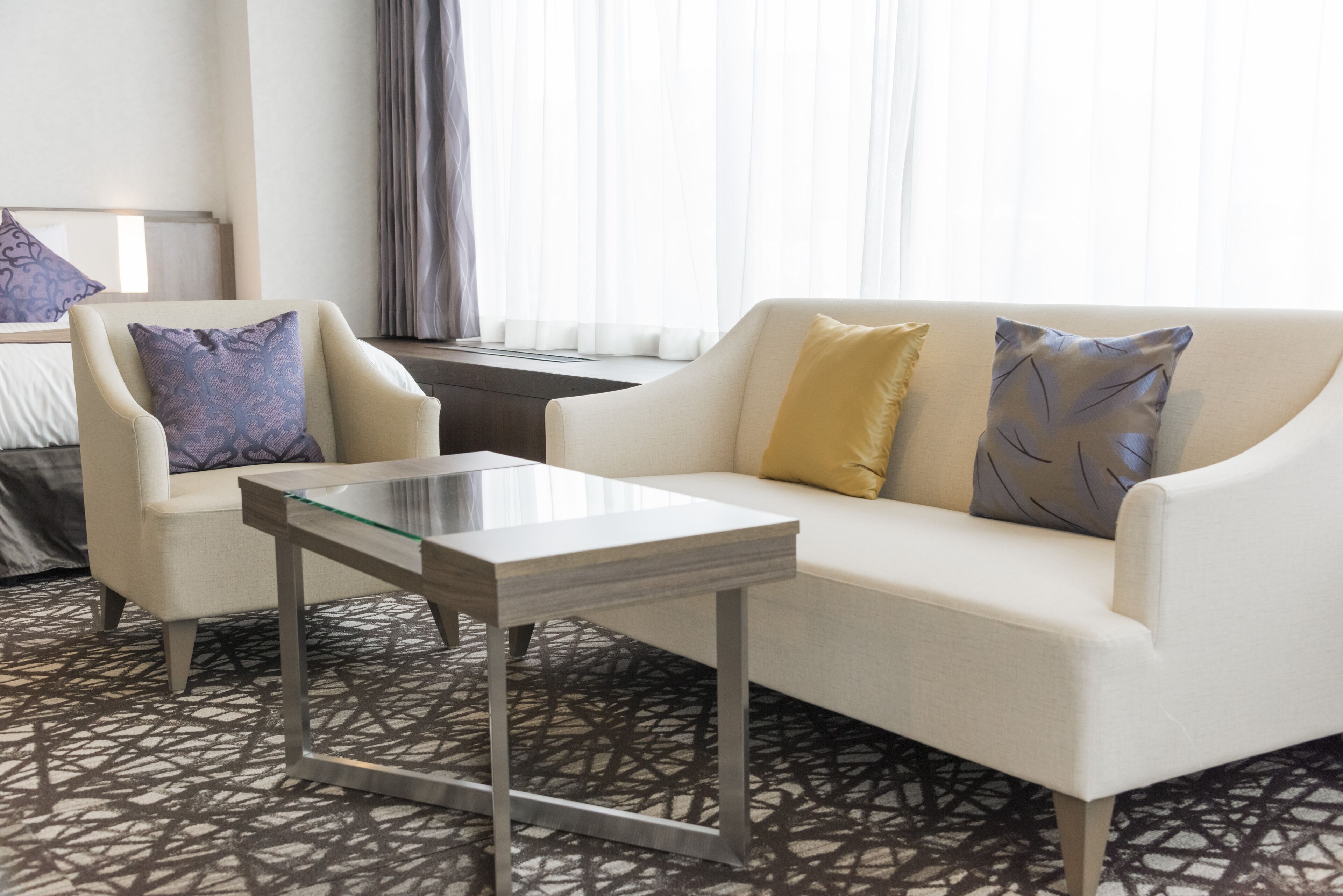 Habitación de hotel con sofá blanco, sillón a juego, mesa de centro de vidrio, alfombra estampada y una ventana con cortinas translúcidas.