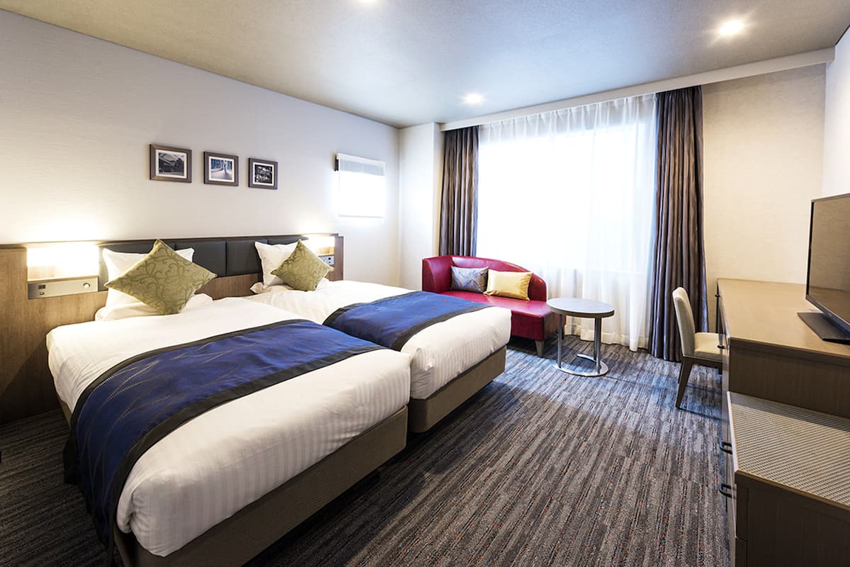 A hotel room with two beds, a desk, a chair, a small table, and a TV.