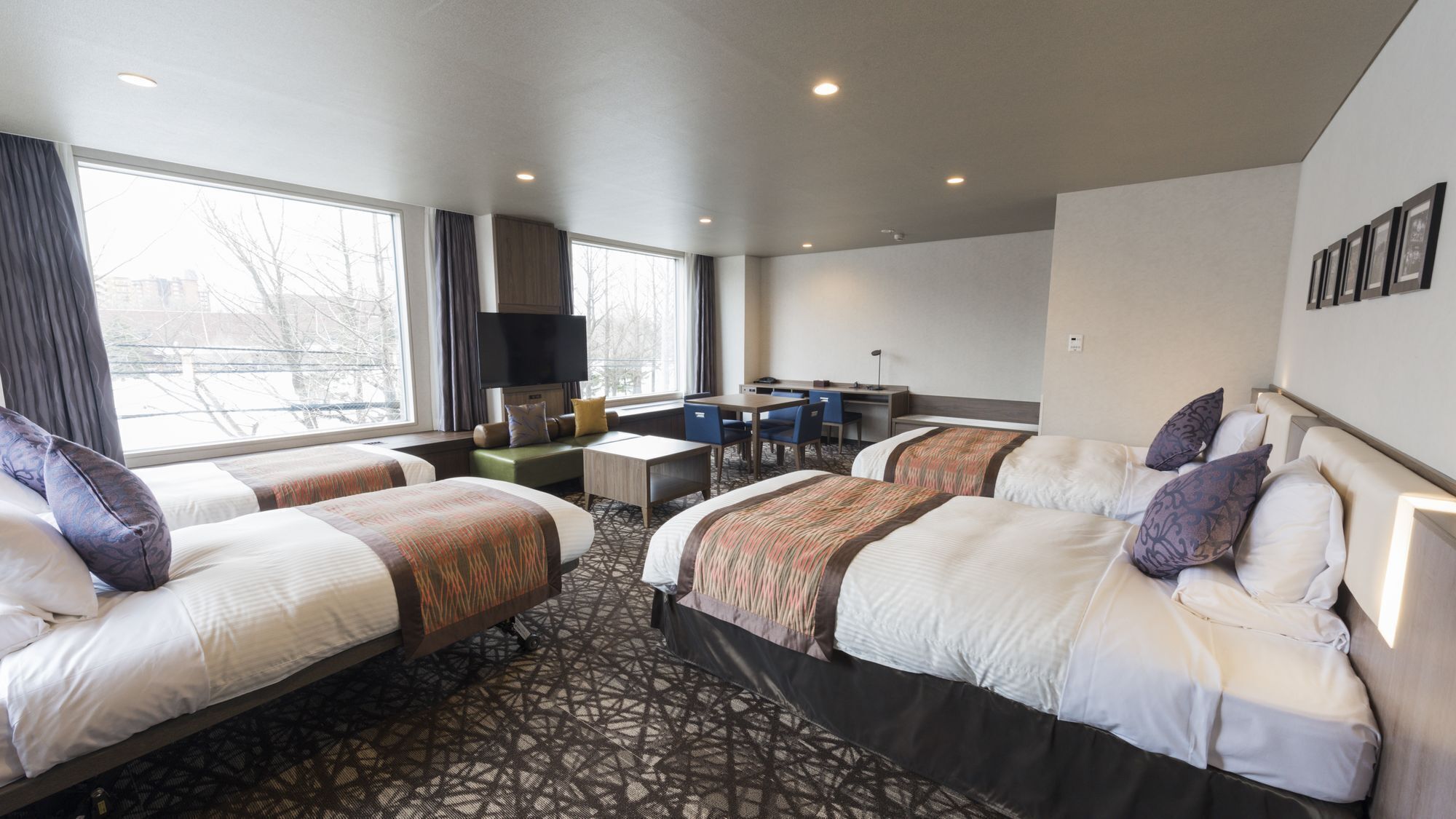 A hotel room with four beds, a desk, a TV, and large windows.