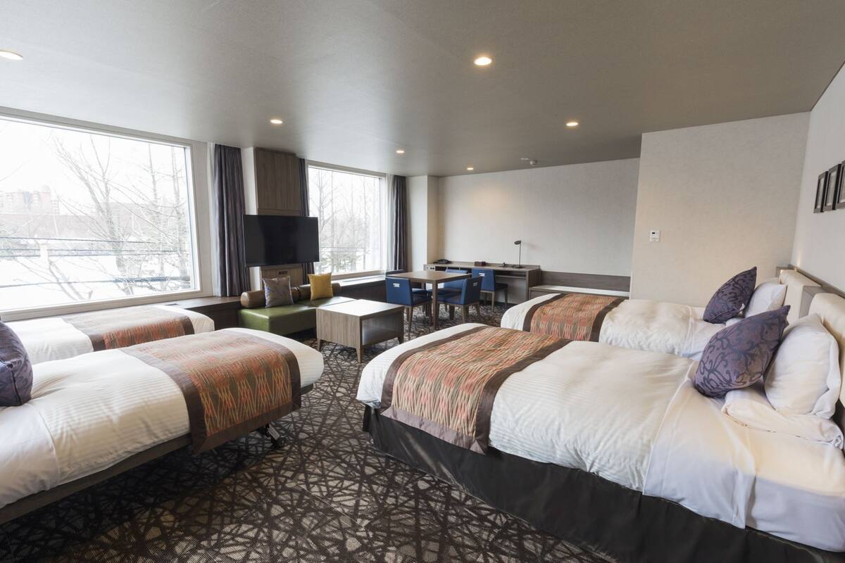 A hotel room with four beds, a desk, a TV, and large windows.