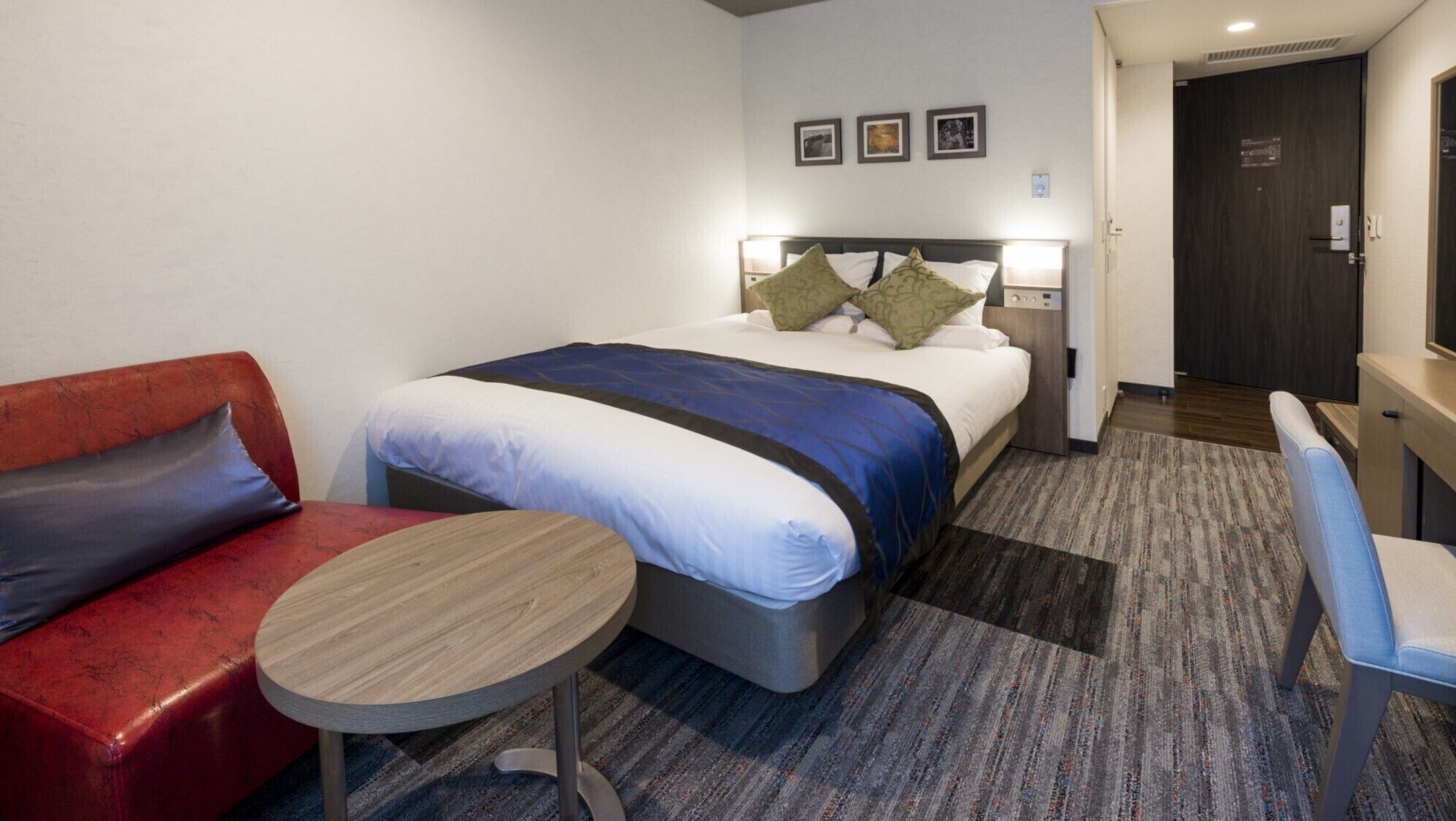 A hotel room with a bed, a red sofa, a small round table, and a door leading to a bathroom.