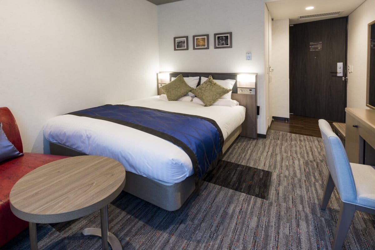 A hotel room with a bed, a red sofa, a small round table, and a door leading to a bathroom.