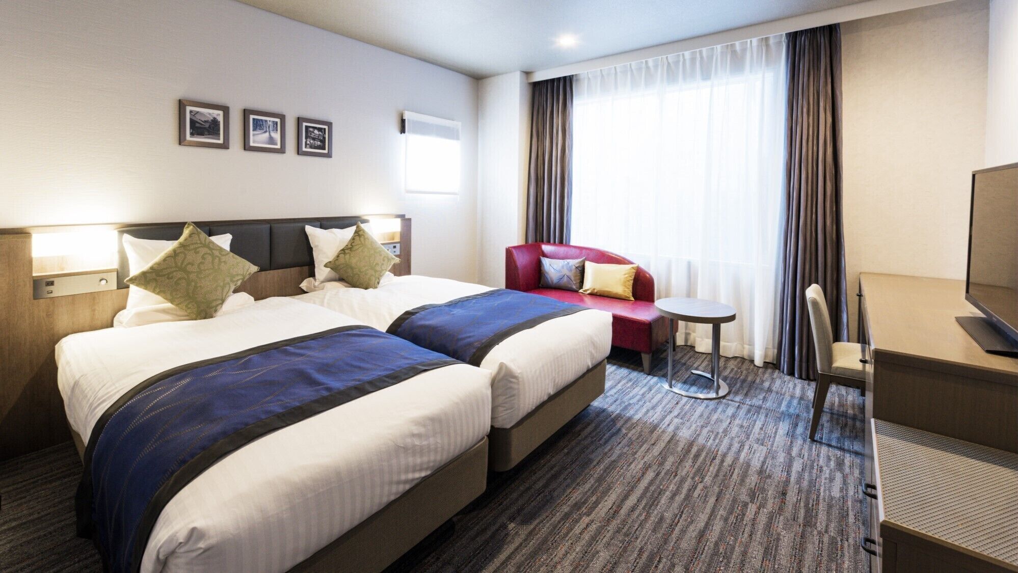 A hotel room with two beds, a red armchair, a small round table, a TV, and three framed pictures on the wall.
