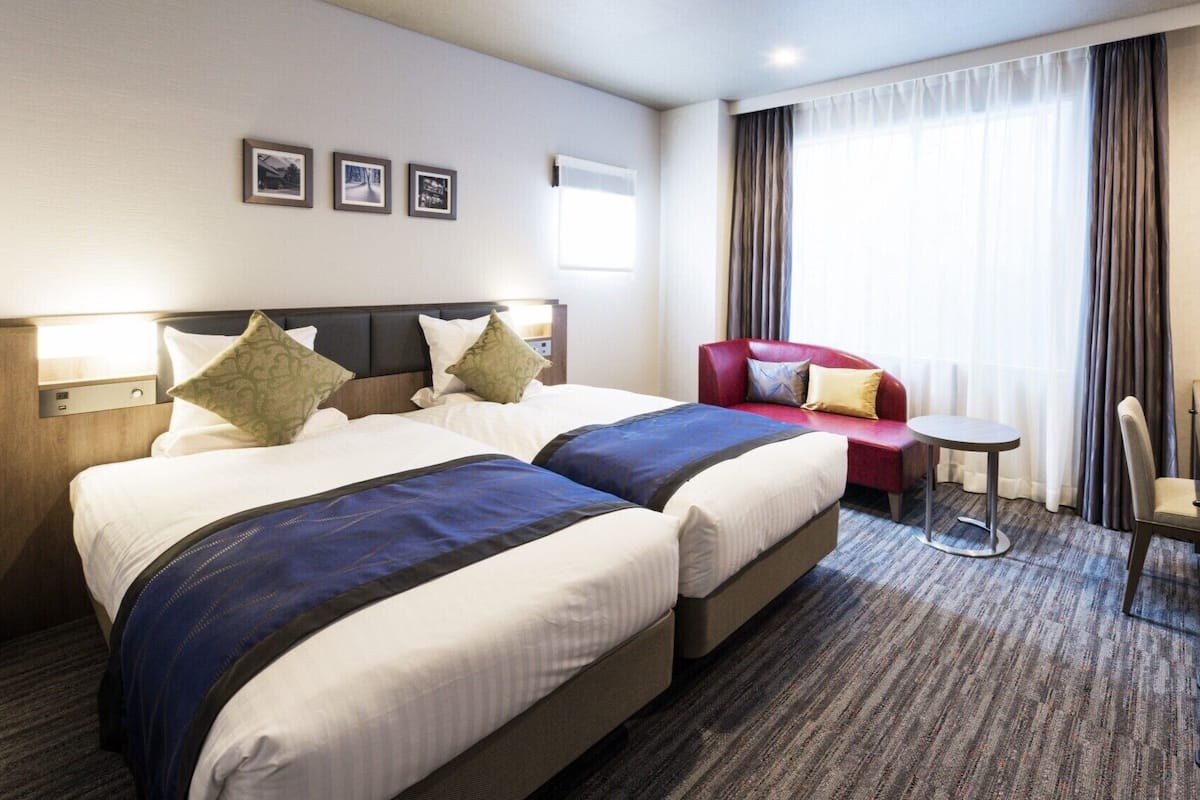 A hotel room with two beds, a red armchair, a small round table, a TV, and three framed pictures on the wall.