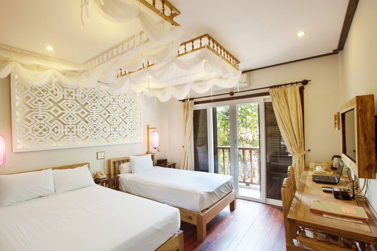 A hotel room with two beds, a wooden desk, a patterned wall, and a balcony with curtains.