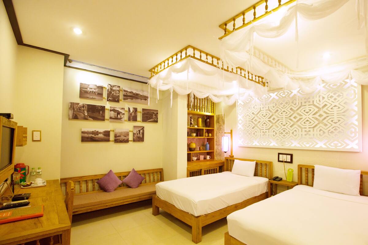 A room with two beds, a bench, a desk with a computer, and a wall with framed pictures.