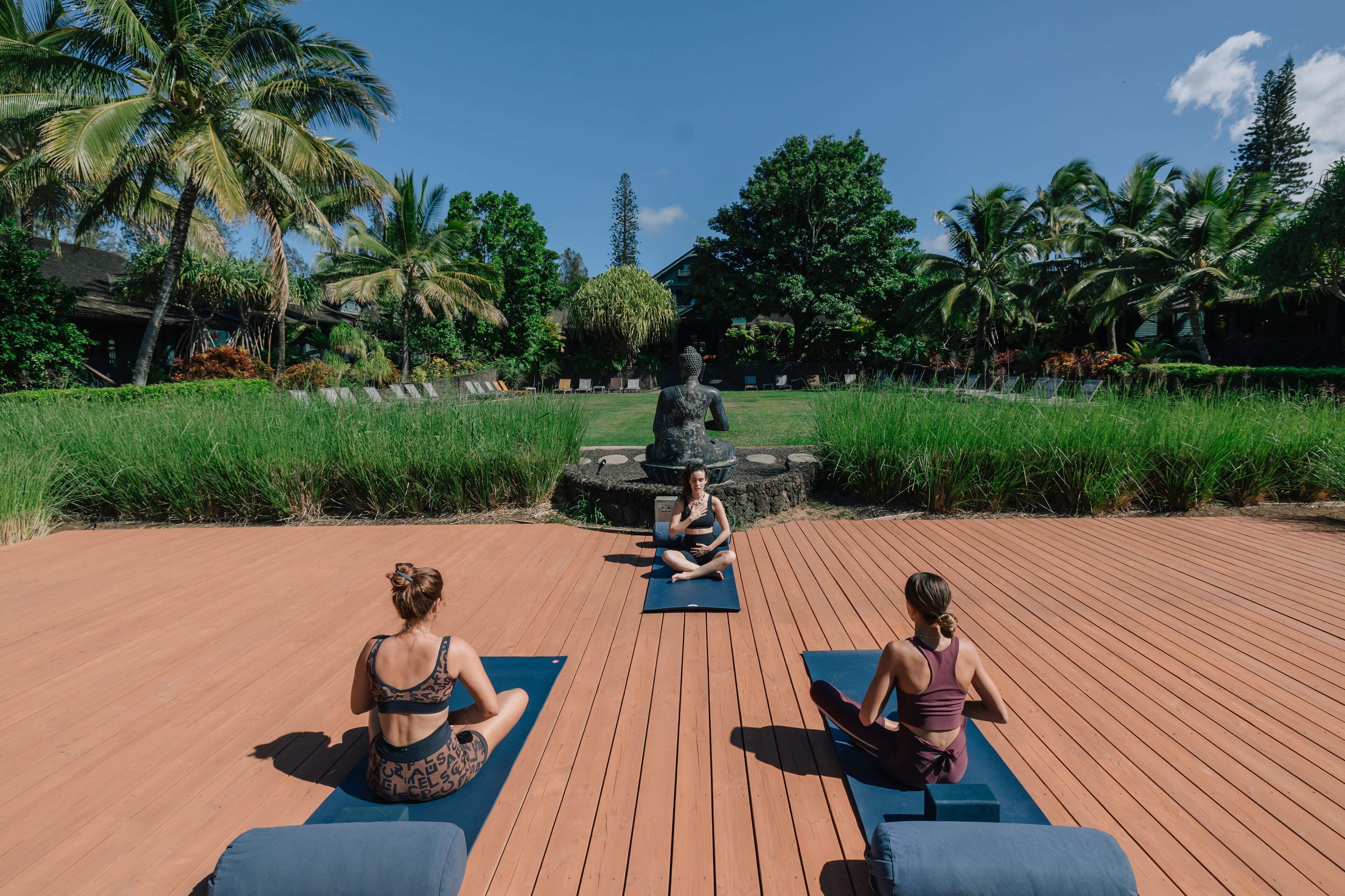 Tres personas sentadas sobre tapetes de yoga en una terraza de madera con una estatua al fondo.