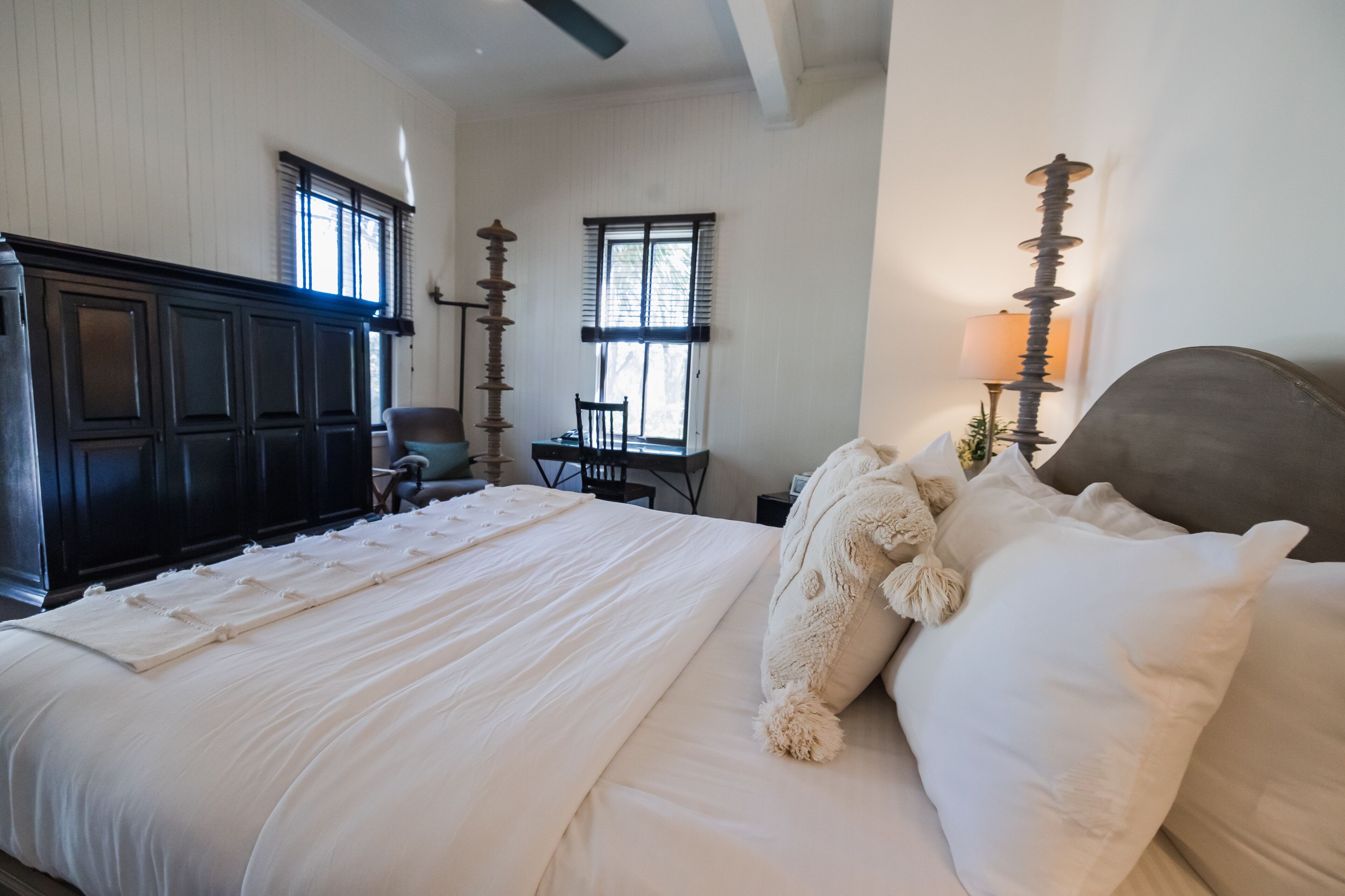 A neatly made bed with white linens and a fluffy pillow, a wooden headboard, and a desk with a chair in a room with white walls and a window with blinds.