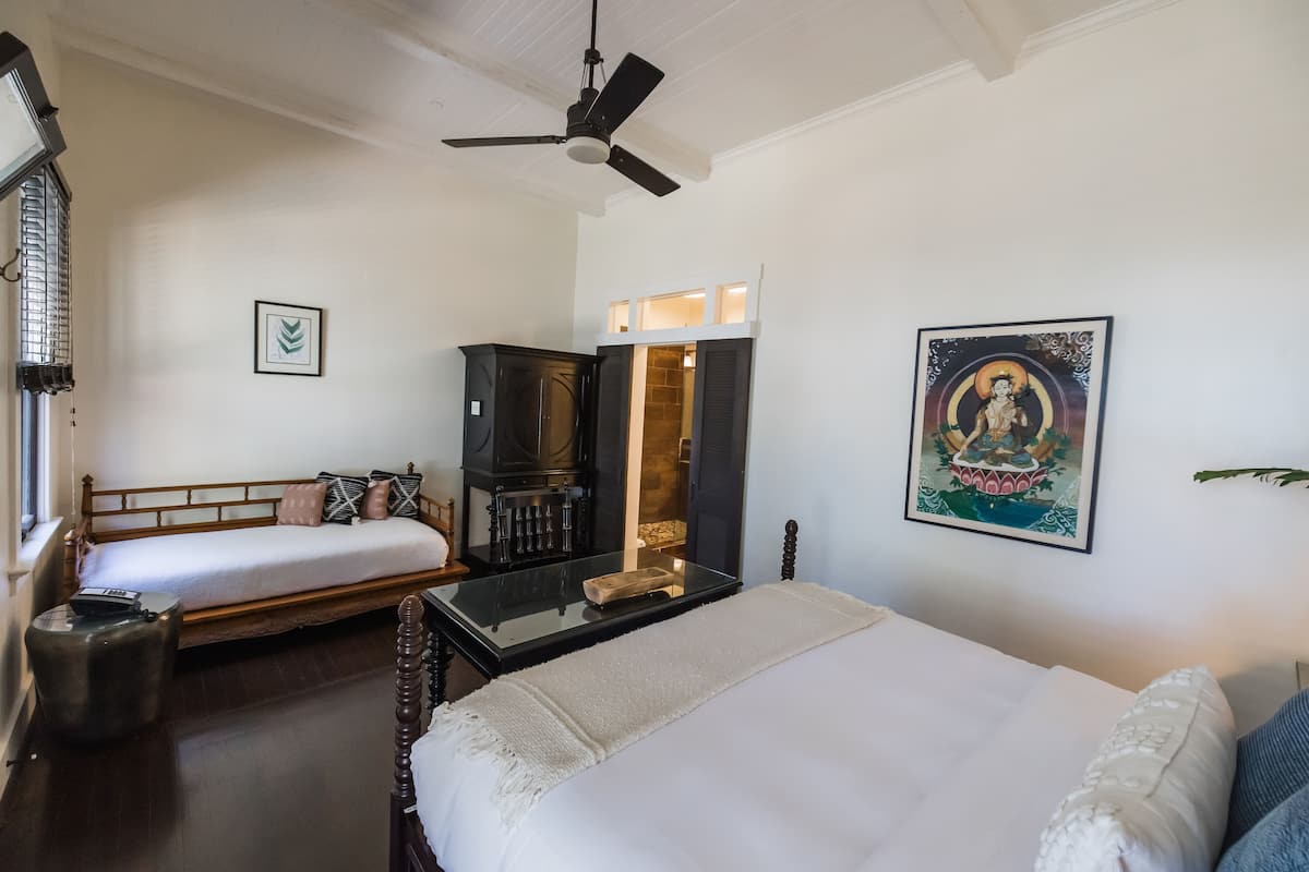 A hotel room with a bed, a desk, a ceiling fan, a window, and a painting on the wall.