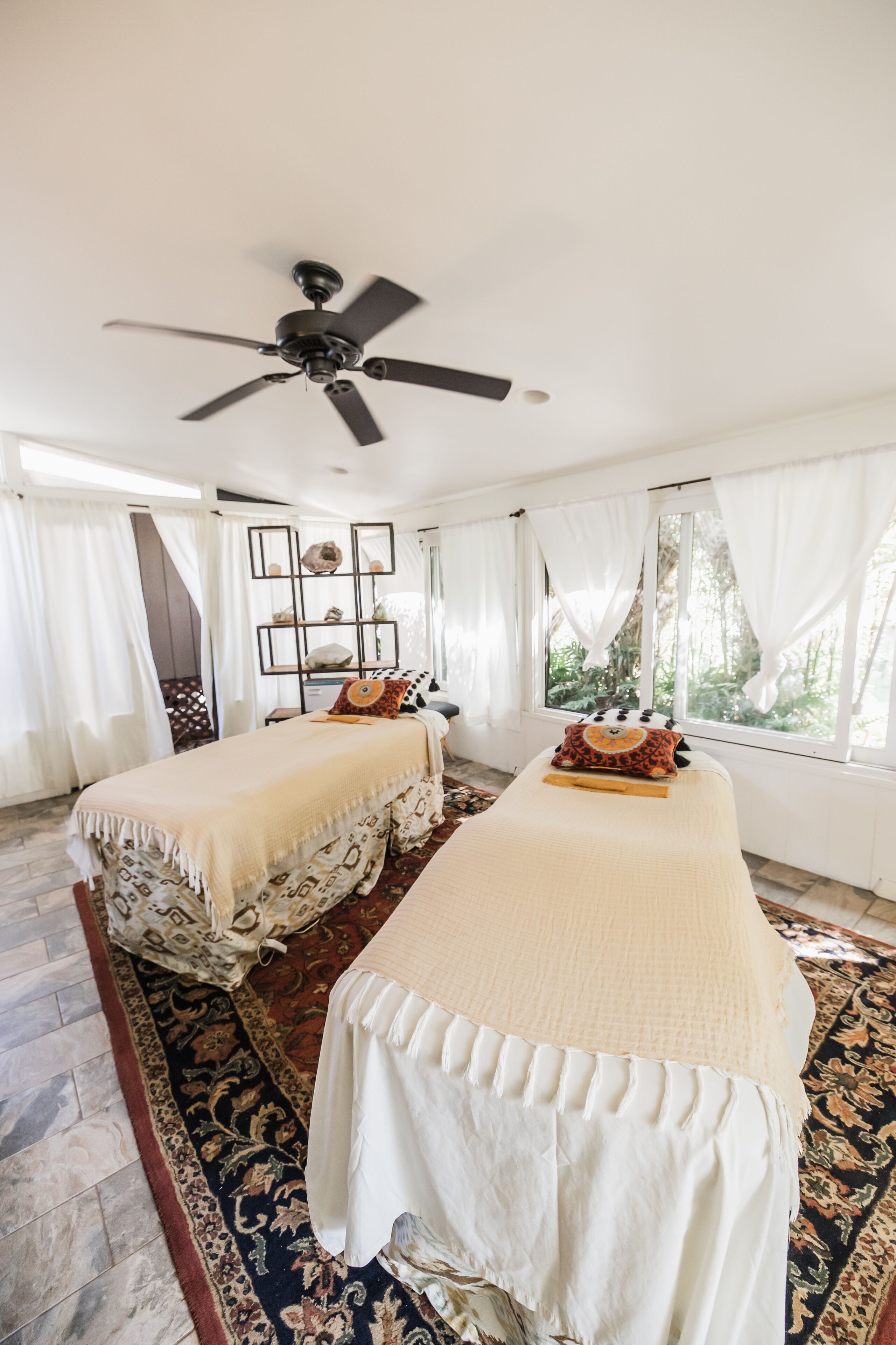 A room with two beds, a ceiling fan, a window with curtains, and a patterned rug.