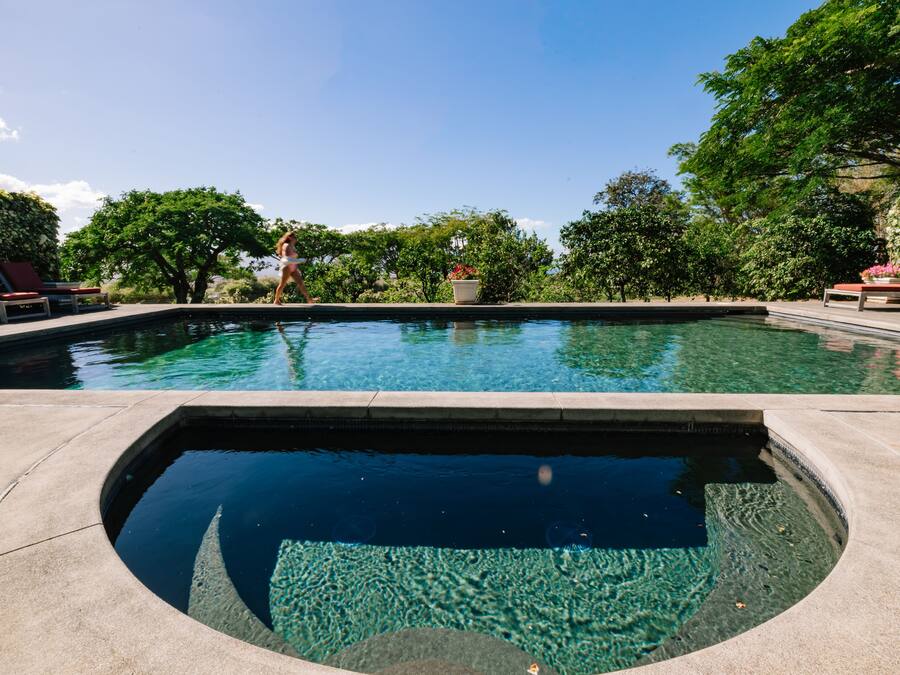 A clear swimming pool with lounge chairs and greenery around.