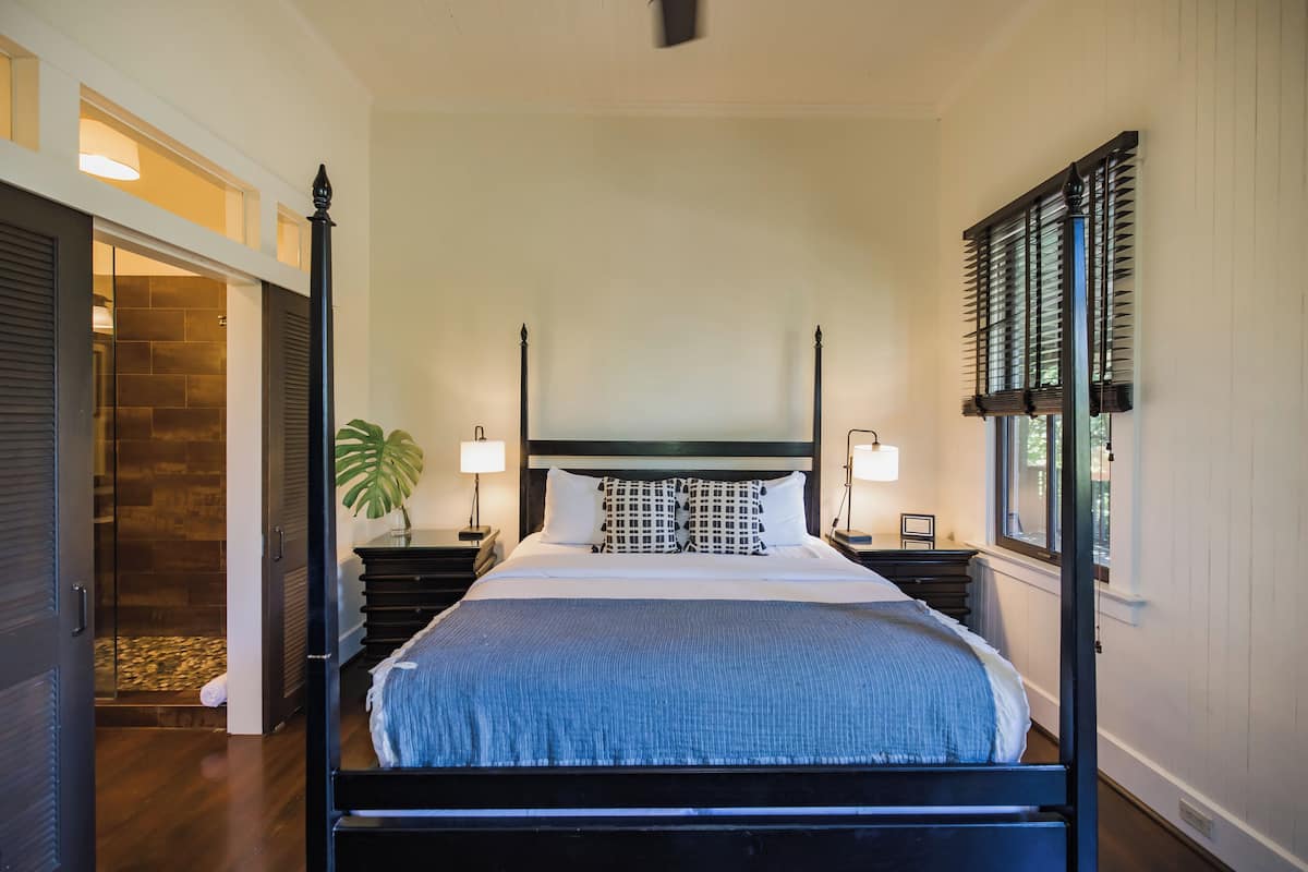 A four-poster bed with a blue bedspread, two bedside tables with lamps, a potted plant, and a view of the outdoors through a window with blinds.
