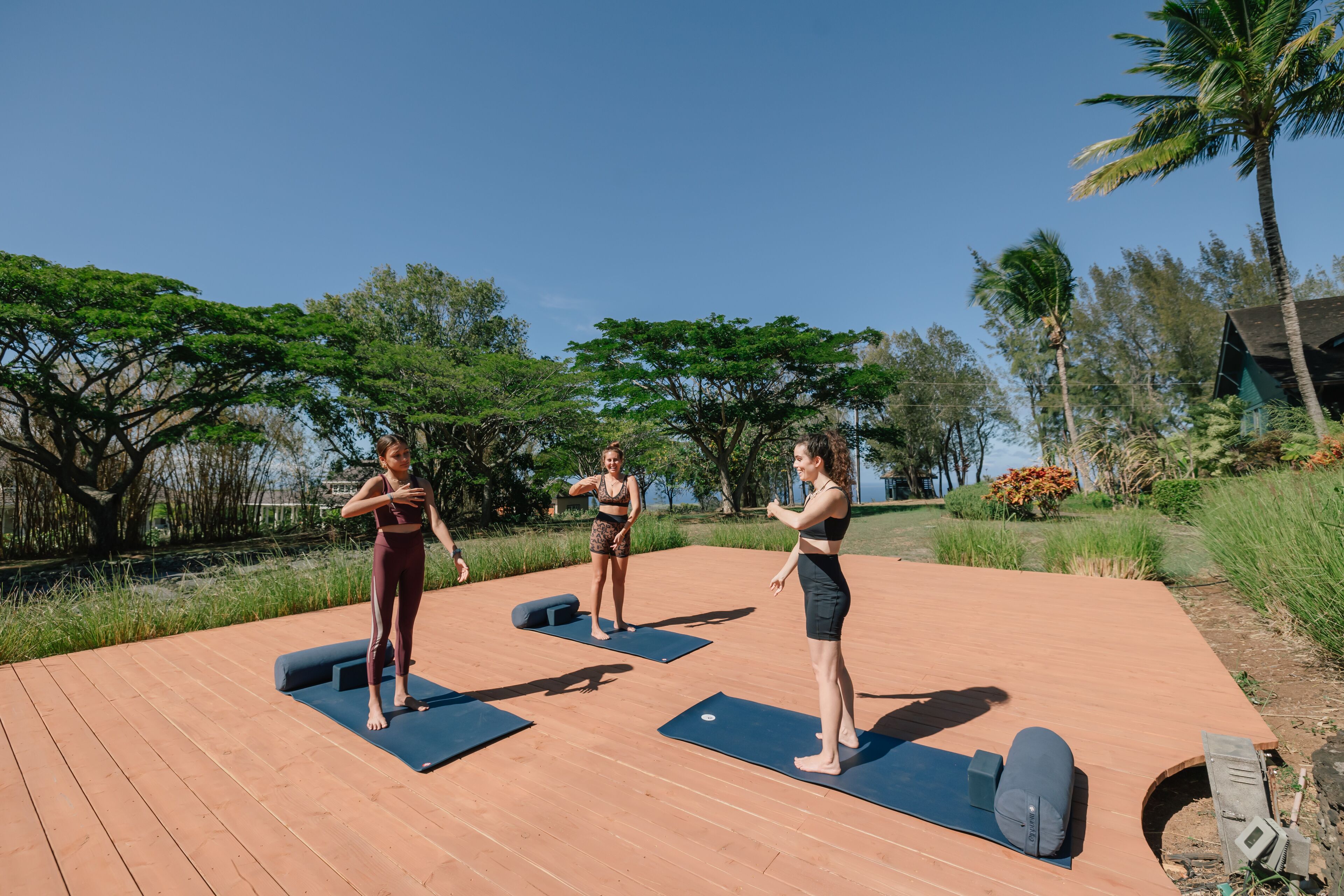 Tres personas sobre colchonetas de yoga en un entorno exterior con una terraza de madera y vegetación.