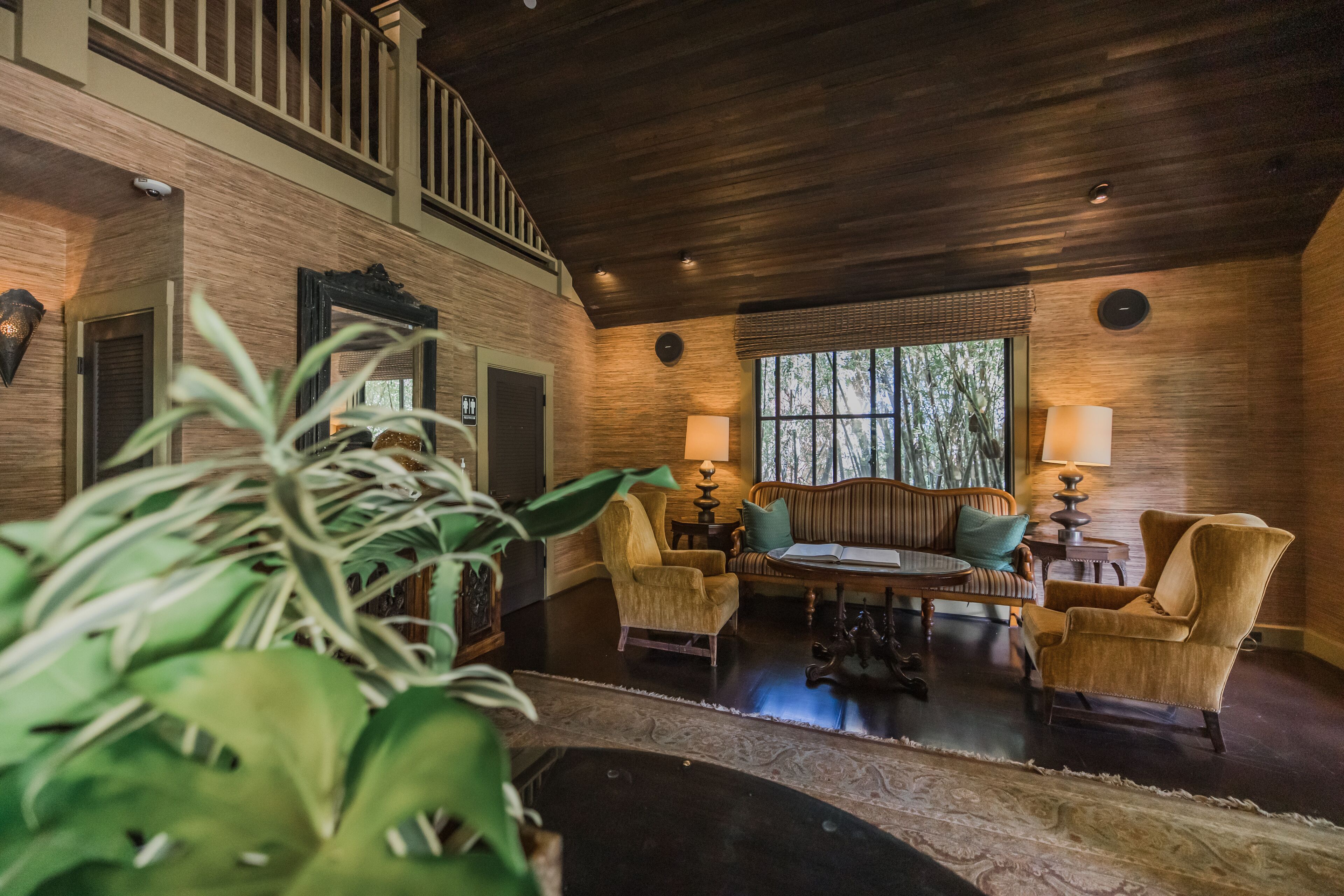 A spacious living room with a high ceiling, wooden flooring, and a large window offering a view of trees. There are several armchairs, a sofa, and a coffee table.