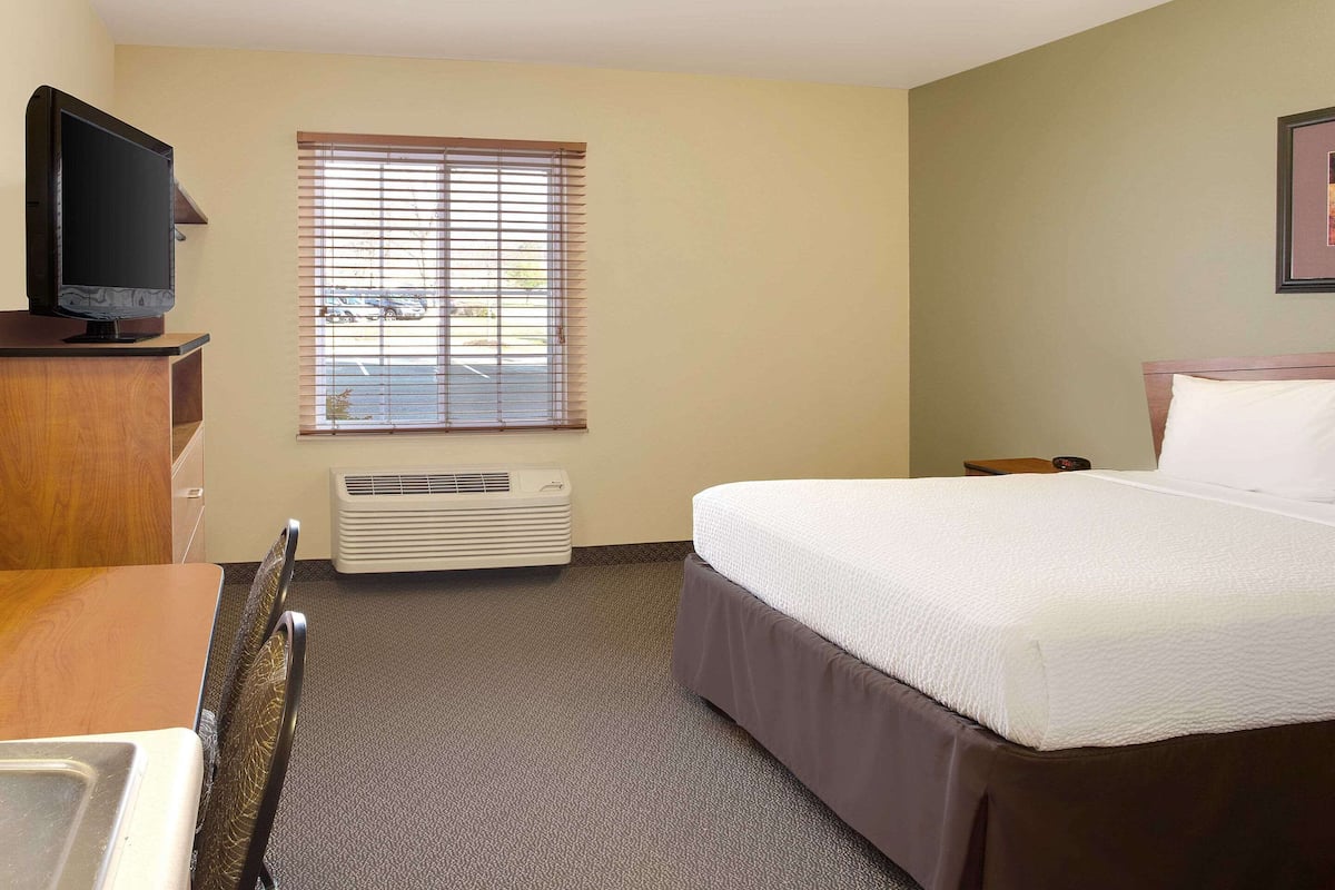 A hotel room with a bed, a television on a dresser, a window with blinds, and a wall-mounted air conditioner.