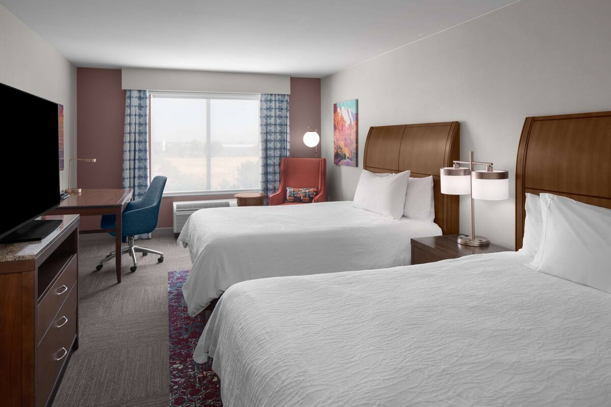 Ein Hotelzimmer mit zwei Betten, einem Schreibtisch mit Fernseher, einem Sessel, einer Lampe und einem Fenster mit Vorhängen.