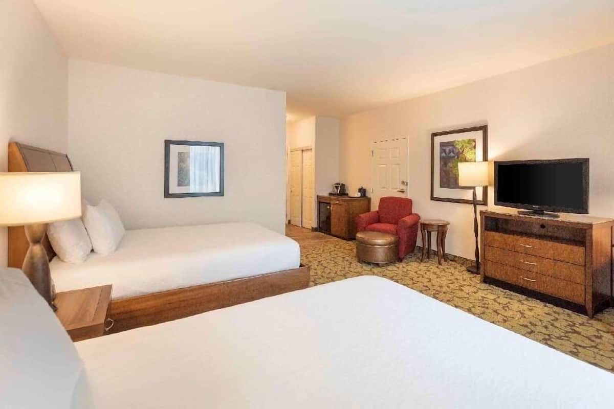 Quarto de hotel com duas camas, uma poltrona vermelha, uma televisão e um aparador de madeira.
