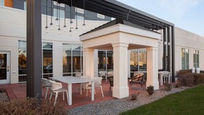An outdoor patio area with tables and chairs, a pergola, and a modern building facade. - Hilton Garden Inn Salt Lake City Airport (Salt Lake City)
