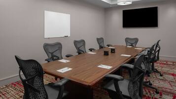 A conference room with a long wooden table, black mesh chairs, a whiteboard, and a flat-screen TV on the wall.