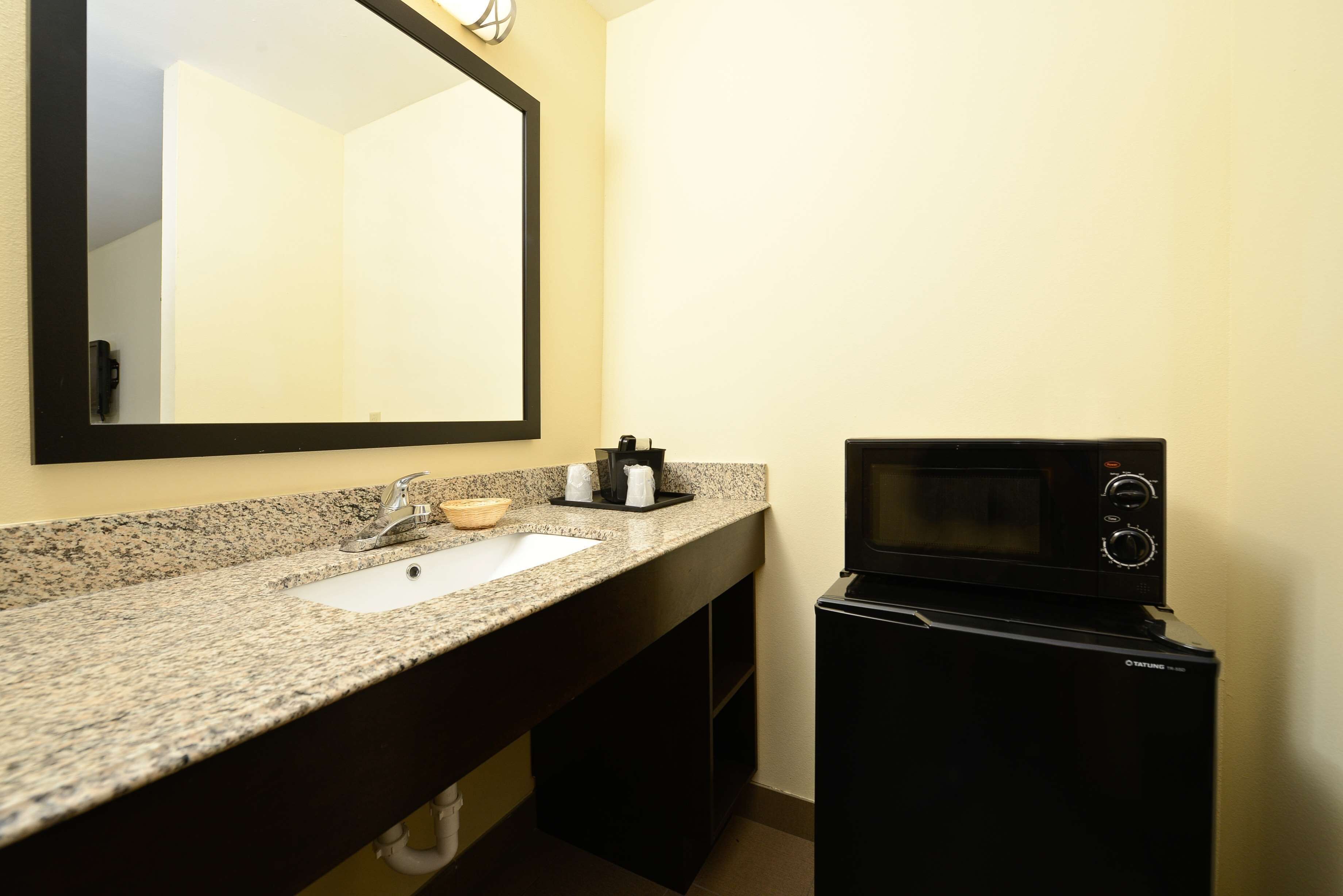 A bathroom with a granite countertop sink, a mirror, a black microwave, and a towel rack.