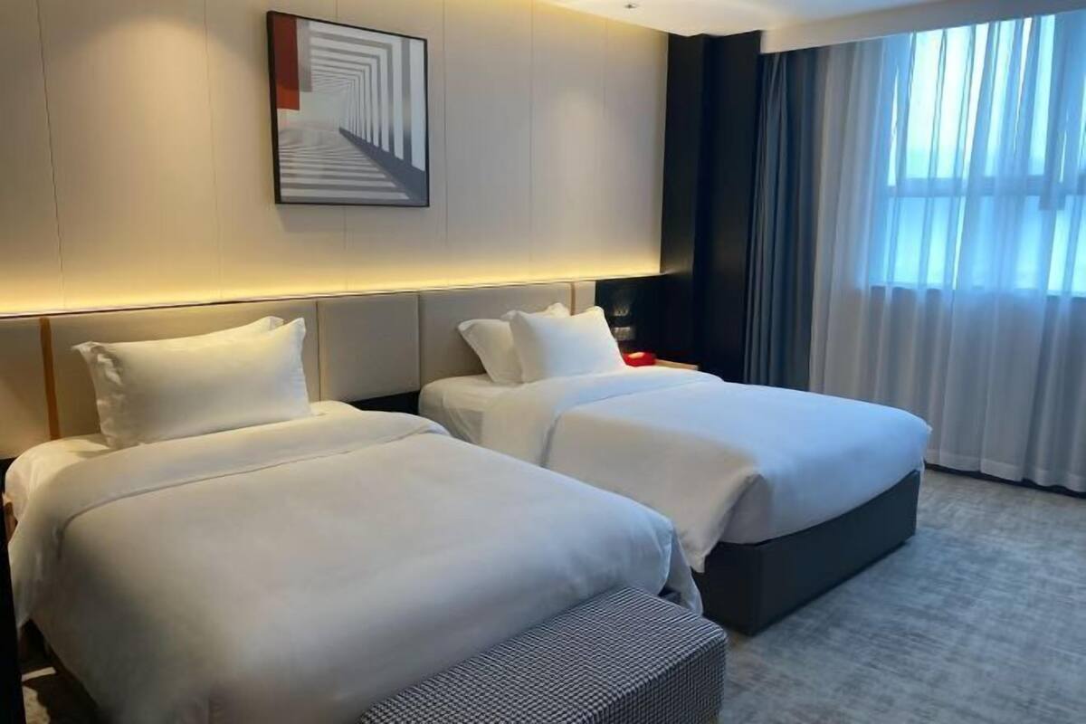 A hotel room with two beds, a modern headboard, a wall-mounted artwork, and a large window with curtains.