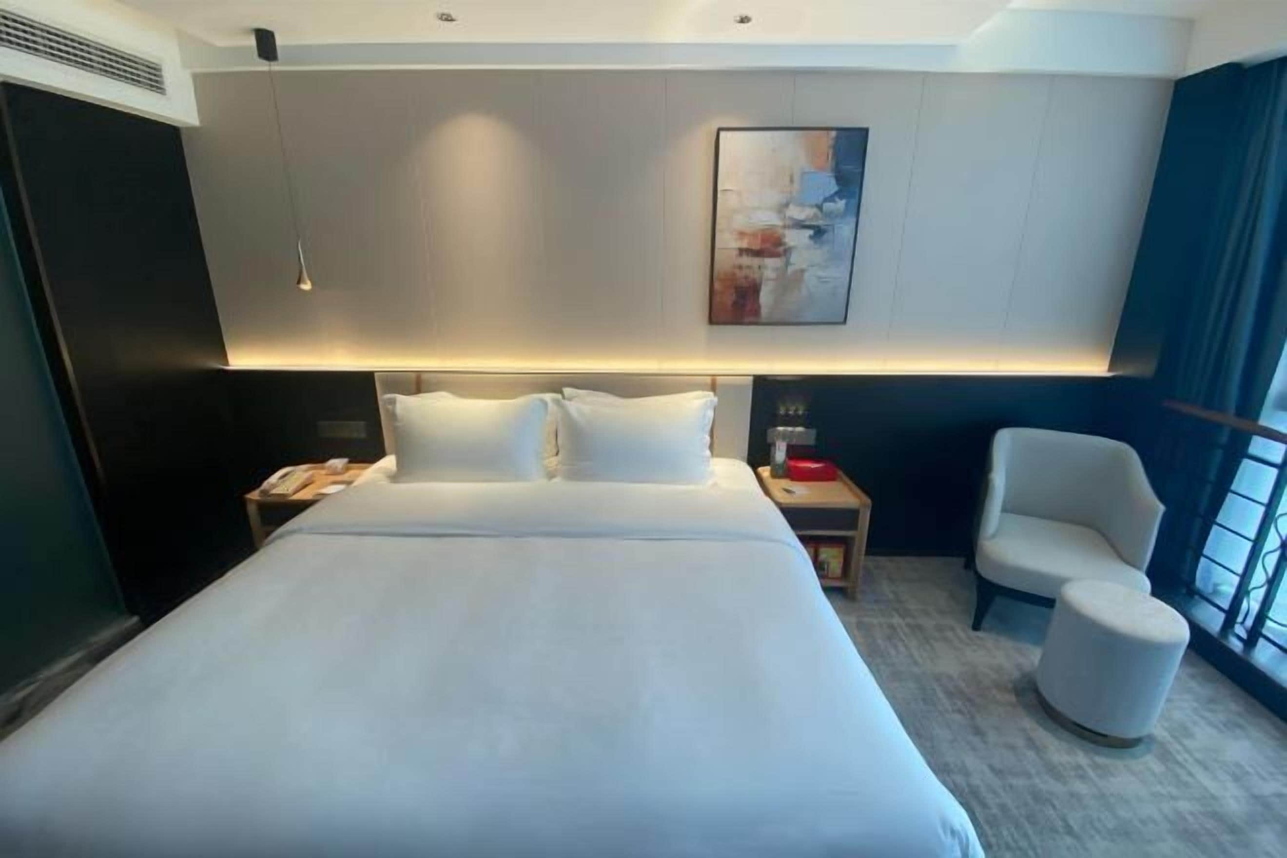 A hotel room with a large bed, a bedside table, a chair, a small ottoman, and a painting on the wall.