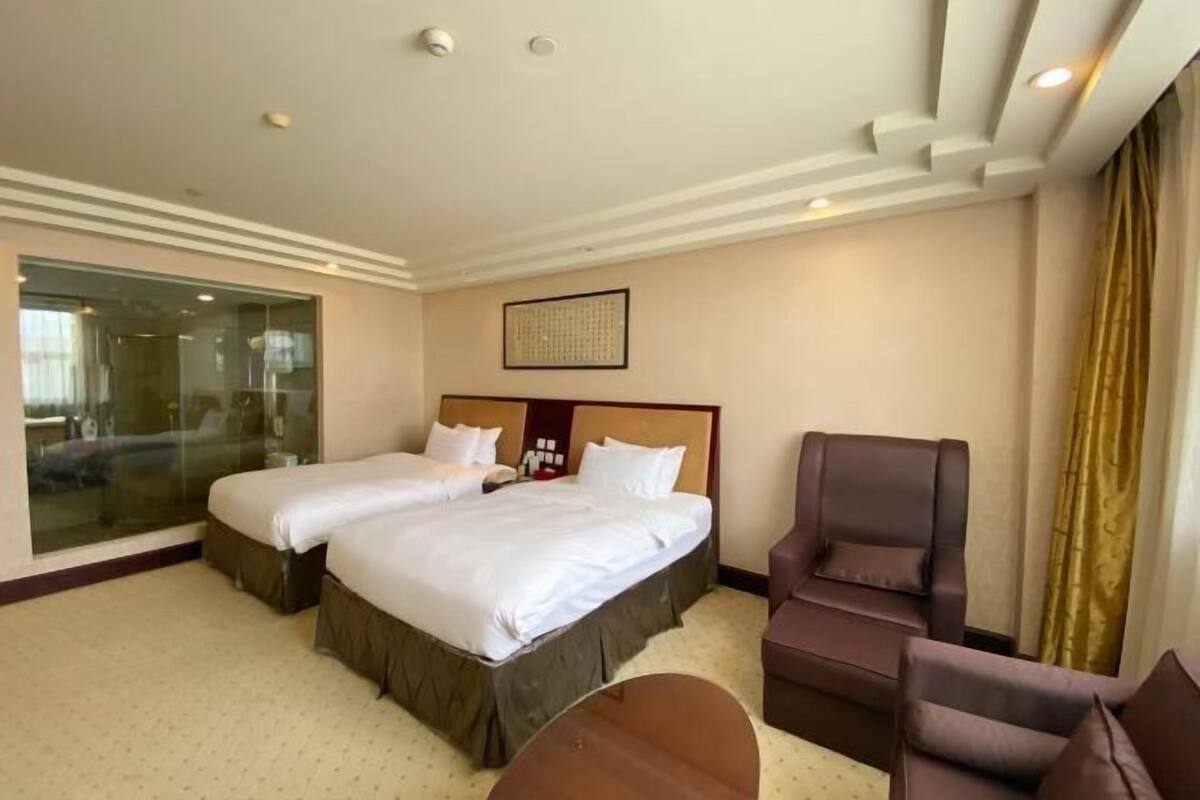 A hotel room with two beds, a seating area with a chair, and a glass partition.