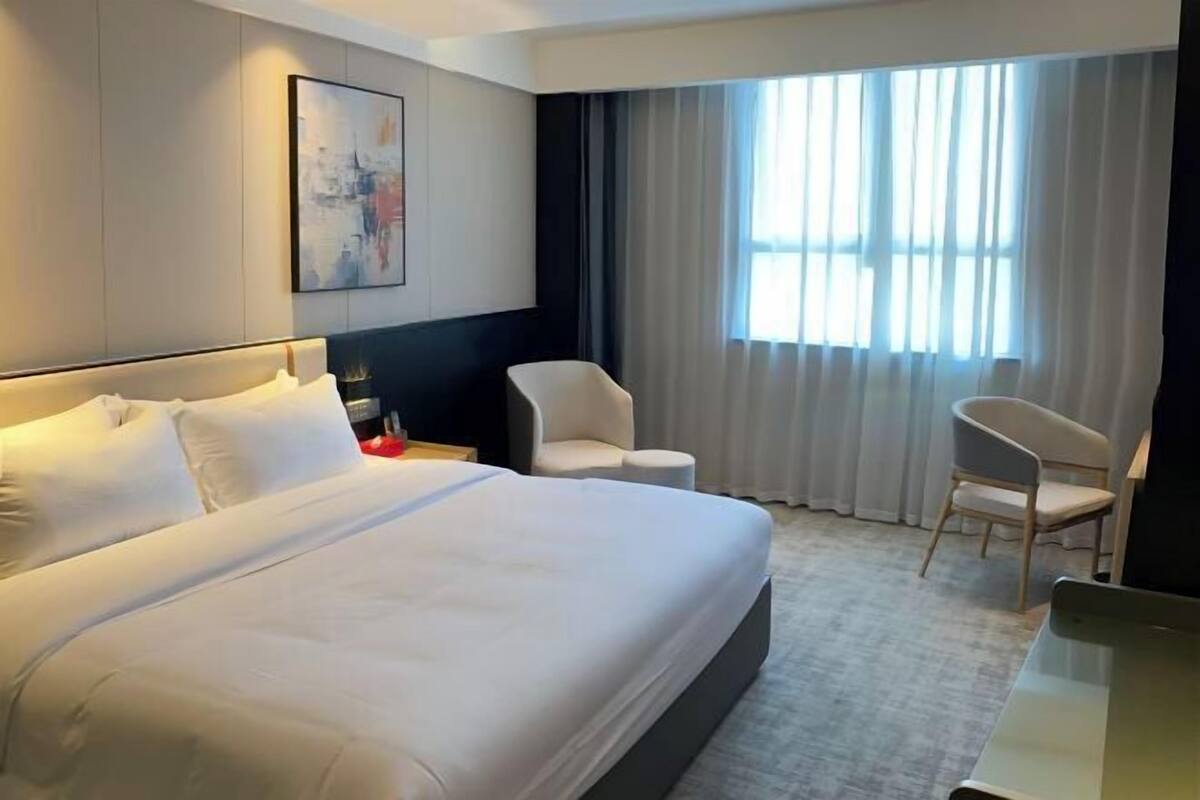 A hotel room with a large bed, two chairs, a desk, a nightstand, and a large window with curtains.