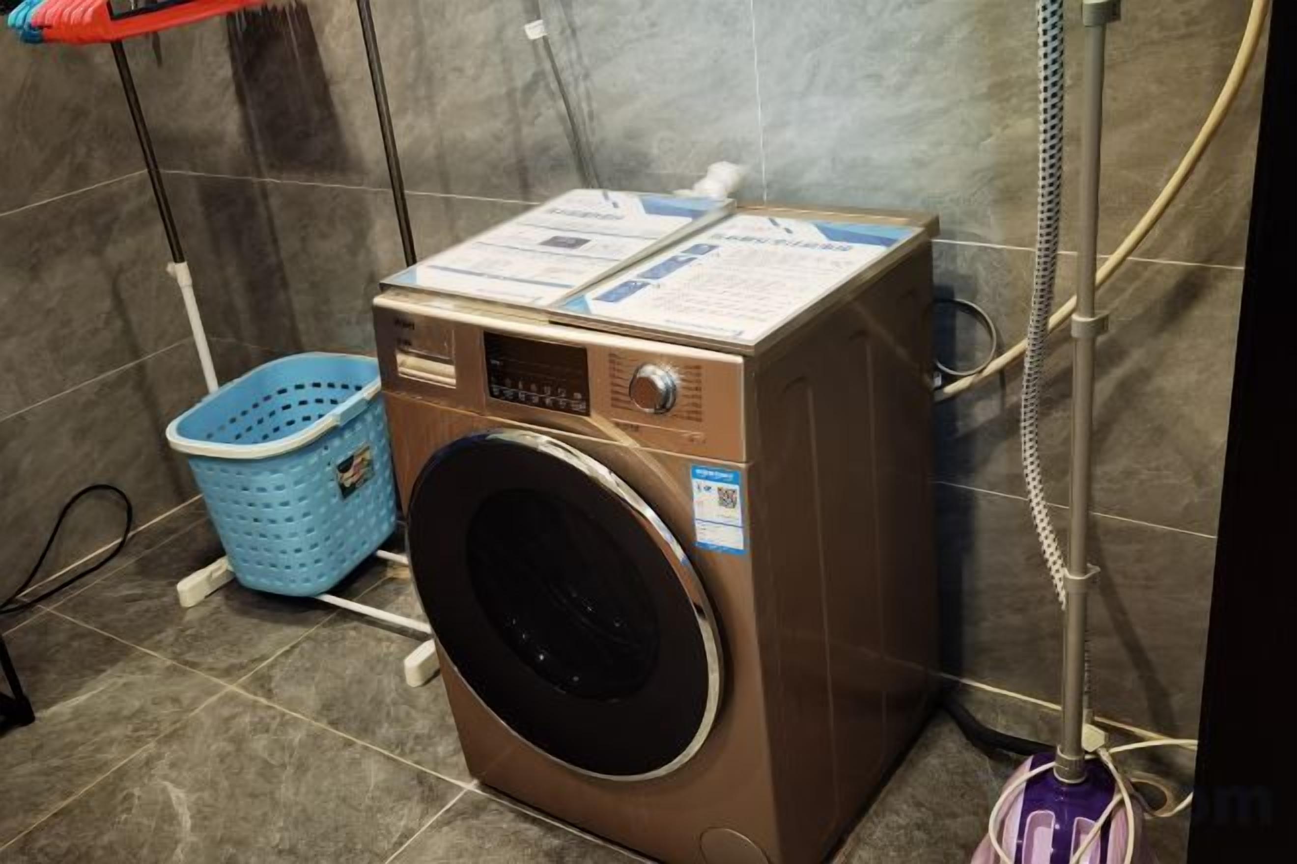 A compact, wall-mounted washing machine with a control panel, a blue laundry basket, and a metal hose.
