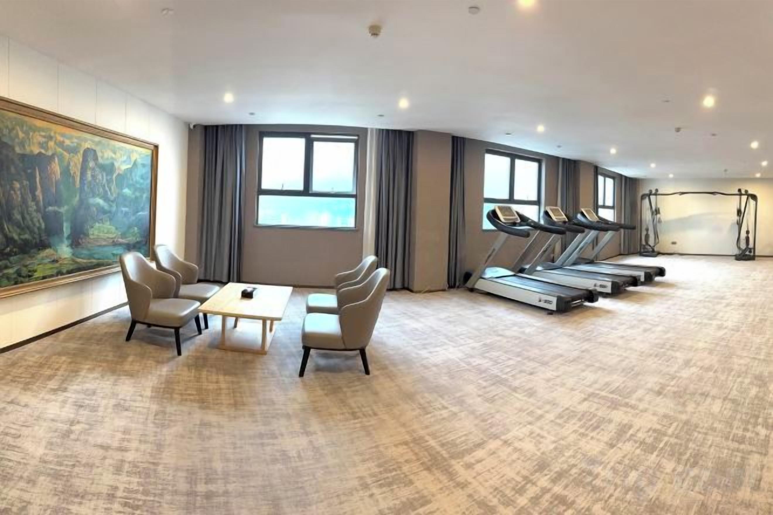 A hotel room with a treadmill, exercise equipment, a large painting, and a seating area with chairs and a table.