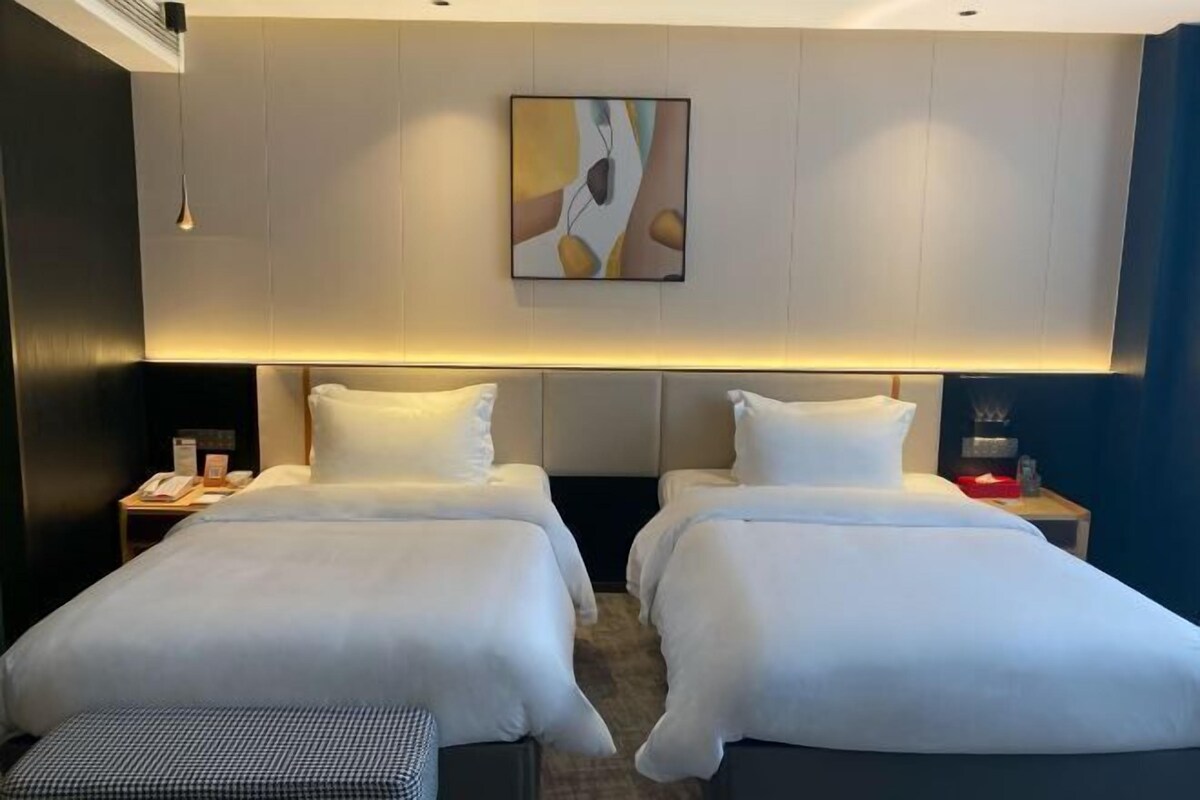 A hotel room with two beds, a bedside table with a lamp, and a wall-mounted artwork.