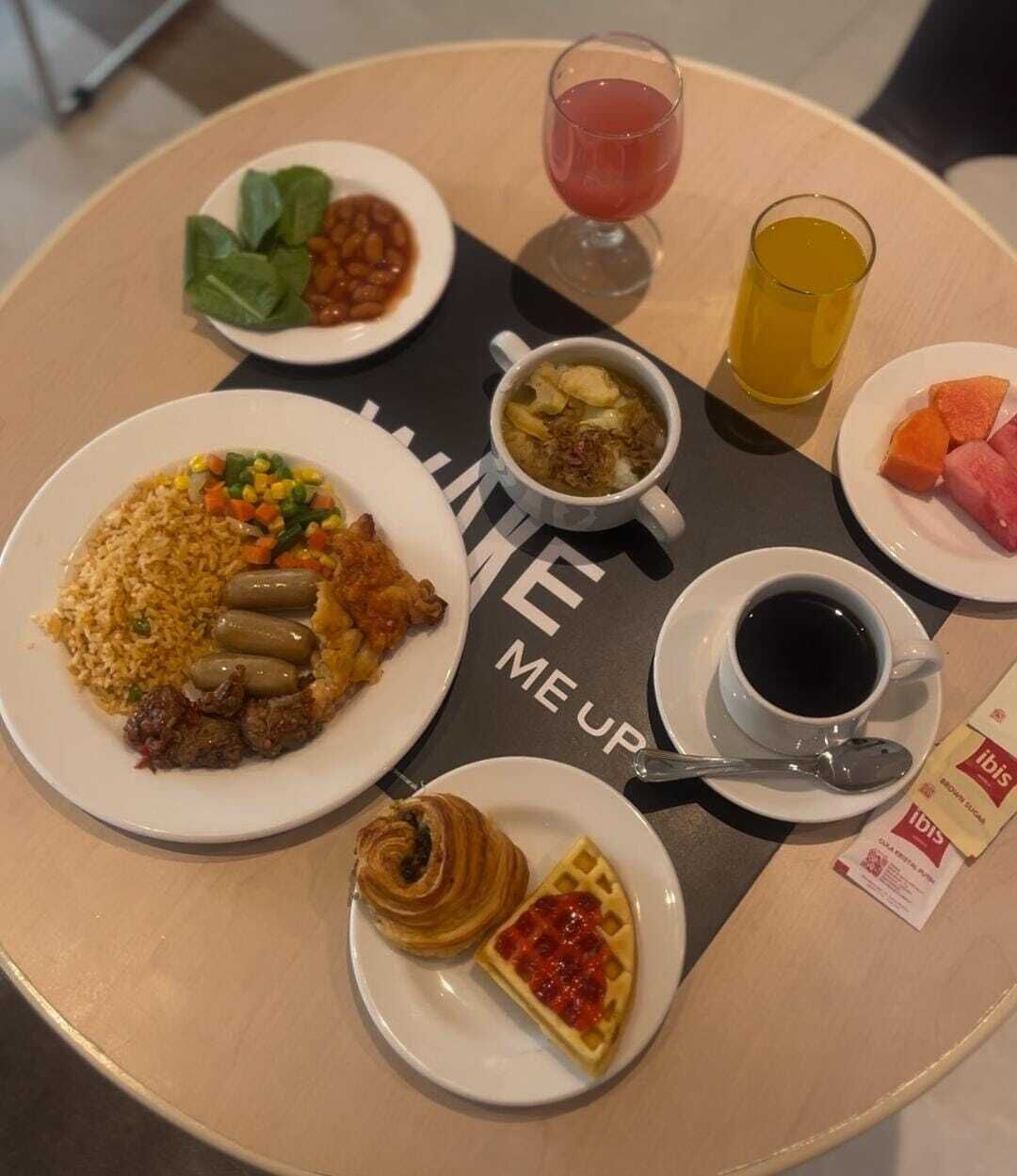 Uma mesa com diversos itens de pequeno-almoço, incluindo arroz, feijão, sopa, sumo, café, croissant e waffle.