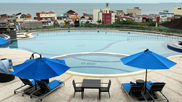 A swimming pool with lounge chairs and umbrellas, a view of the sea, and buildings in the background. - ibis Balikpapan (Balikpapan)