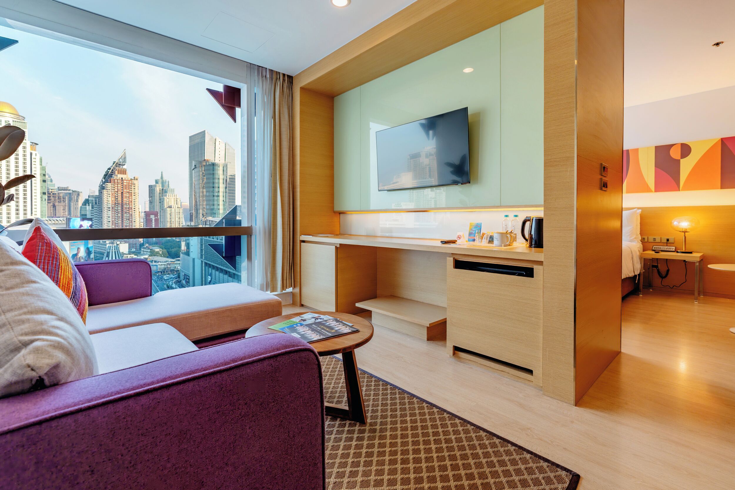 A modern hotel room with a city view, a sofa, a small table, a flat-screen TV, and a kitchenette.