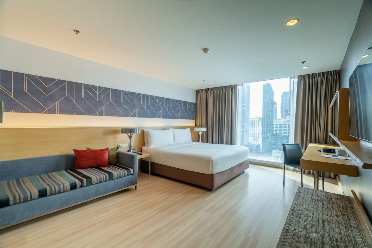 A modern hotel room with a large bed, a sofa, a desk, and a view of the city skyline.