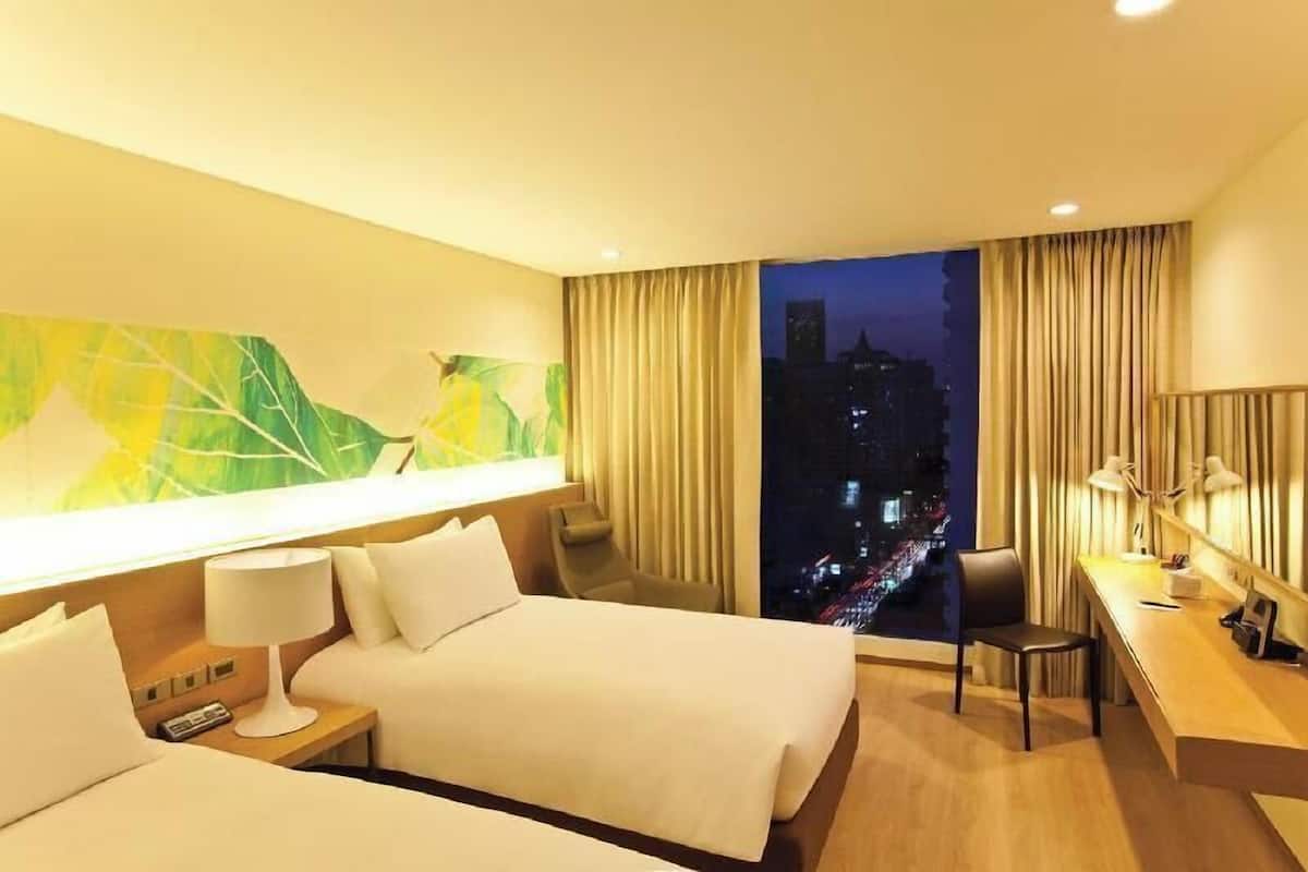 A hotel room with a bed, a desk, a chair, a nightstand, a lamp, and a large window with curtains.