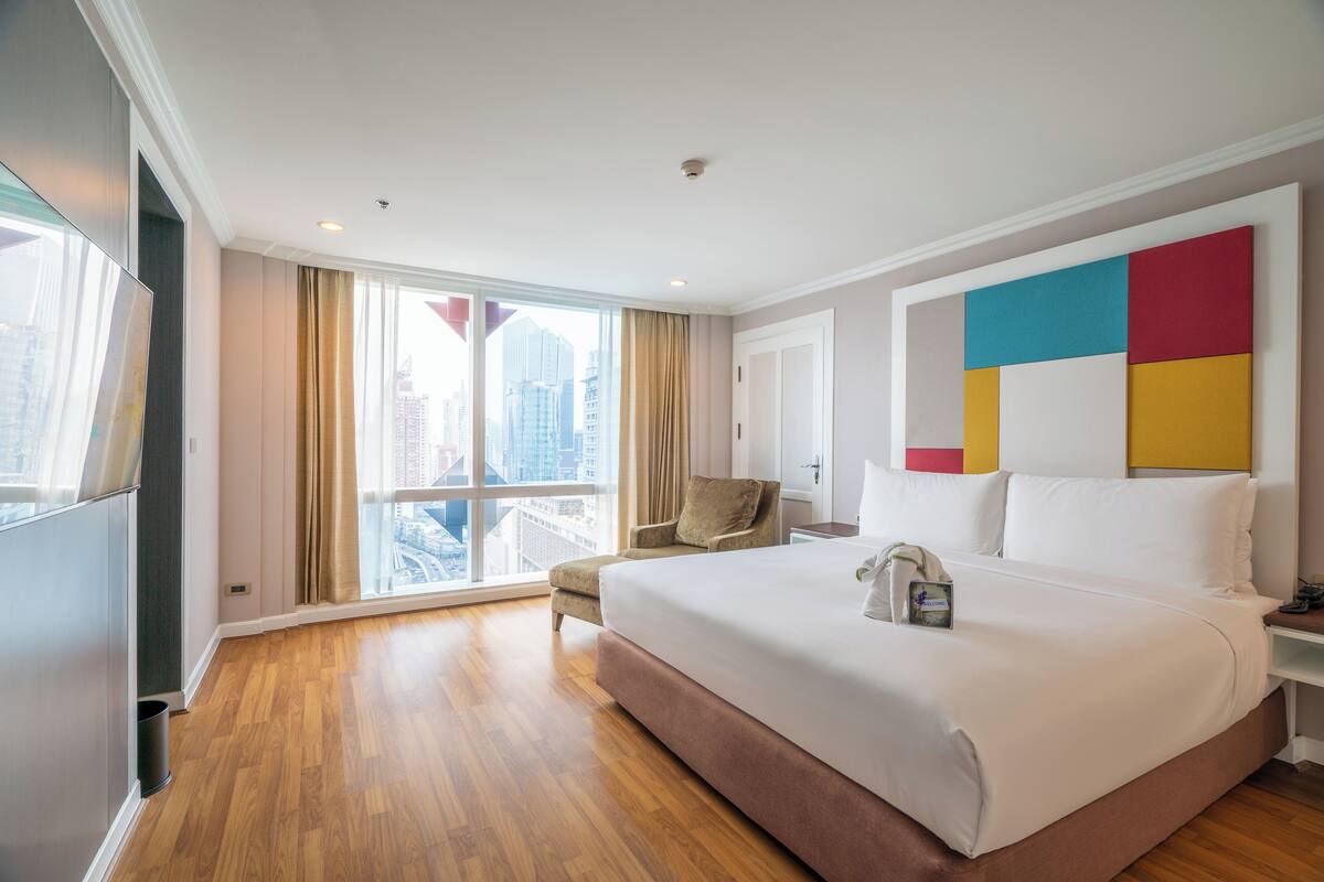 A hotel room with a large bed, a chair, a desk, and a view of the city through large windows.