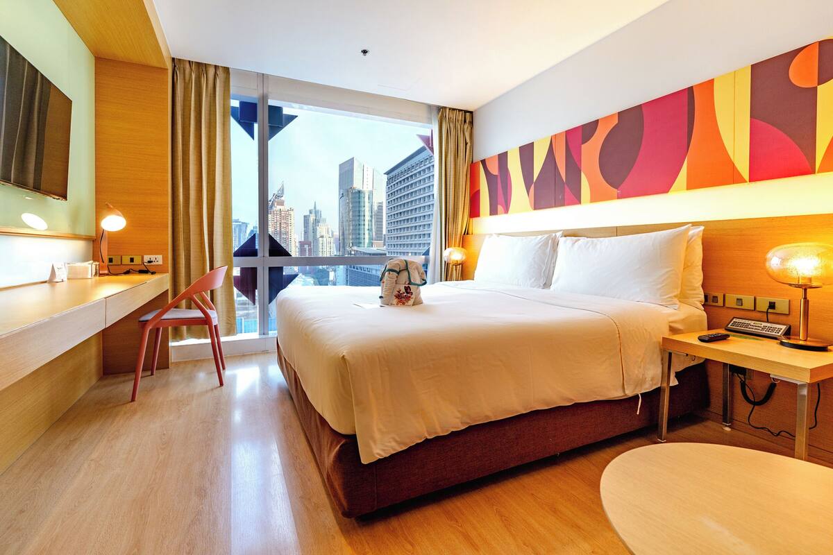 A hotel room with a bed, desk, chair, and a view of the cityscape.