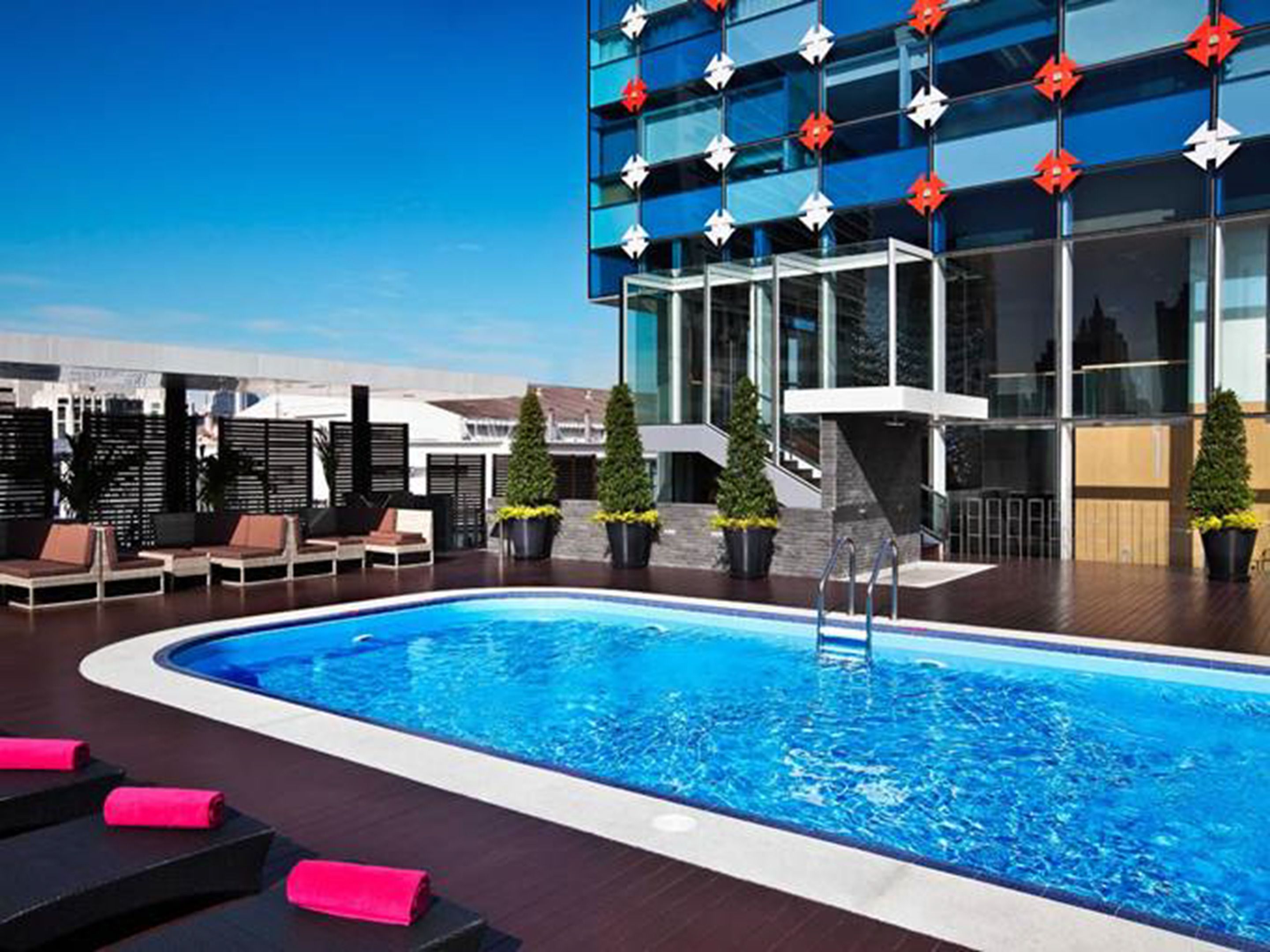 Un área de piscina en la azotea con sillones de descanso, una piscina y un edificio moderno con detalles de vidrio y toques rojos.
