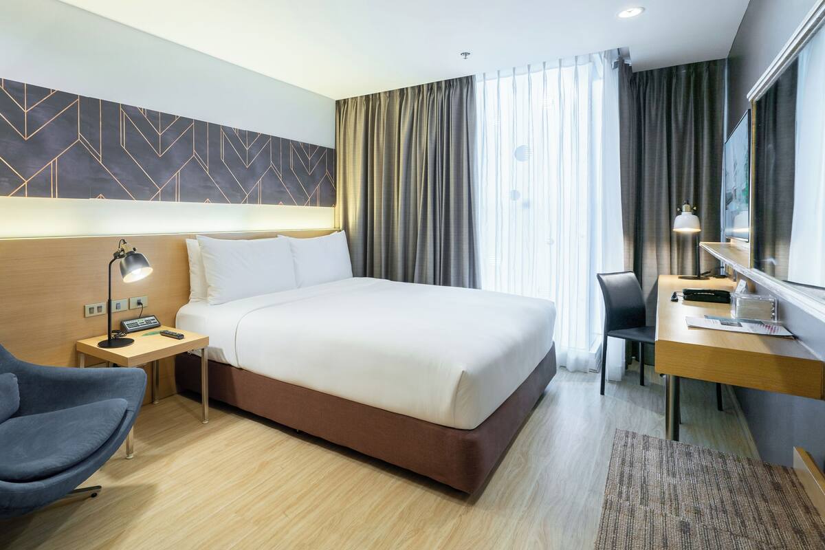 A hotel room with a bed, a desk with a lamp, a chair, and a headboard with a geometric pattern.