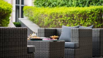 Wicker furniture with cushions, a table with a teapot and plates, and a neatly trimmed hedge in the background.