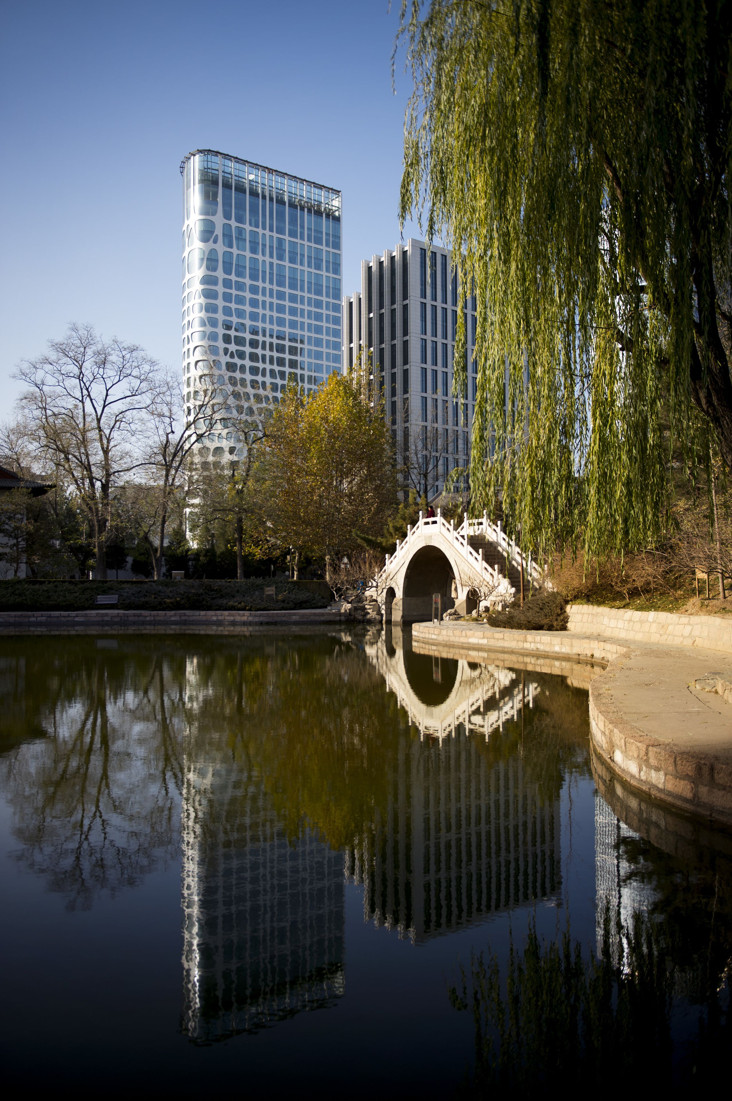A modern building with a unique glass facade, a traditional stone bridge over a reflective water body, and surrounding trees.