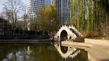 Un edificio moderno con una fachada de vidrio única, un puente de piedra tradicional sobre una superficie acuática reflectante y árboles circundantes.