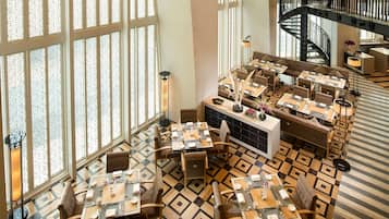 Un vestíbulo de hotel con una zona de estar central, varias mesas de comedor, una escalera y amplias ventanas con un diseño geométrico en el suelo.