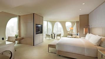 A modern hotel room with a large bed, a TV, a desk, and a seating area with a view of the city.